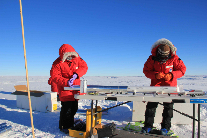 Zdjęcie przedstawia dwoje naukowców w czerwonych kurtkach, zabezpieczających zamknięty w specjalnym pojemniku podłużny rdzeń lodowy. W tle lodowy krajobraz.