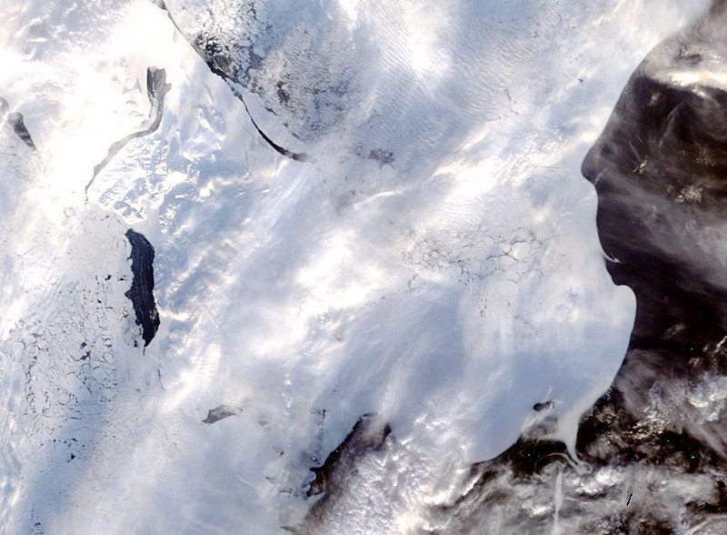 Zdjęcie przedstawia widzianą z góry, pękającą skorupę lodową (w szczelinach widoczne jest czarne morze), częściowo przesłoniętą chmurami.