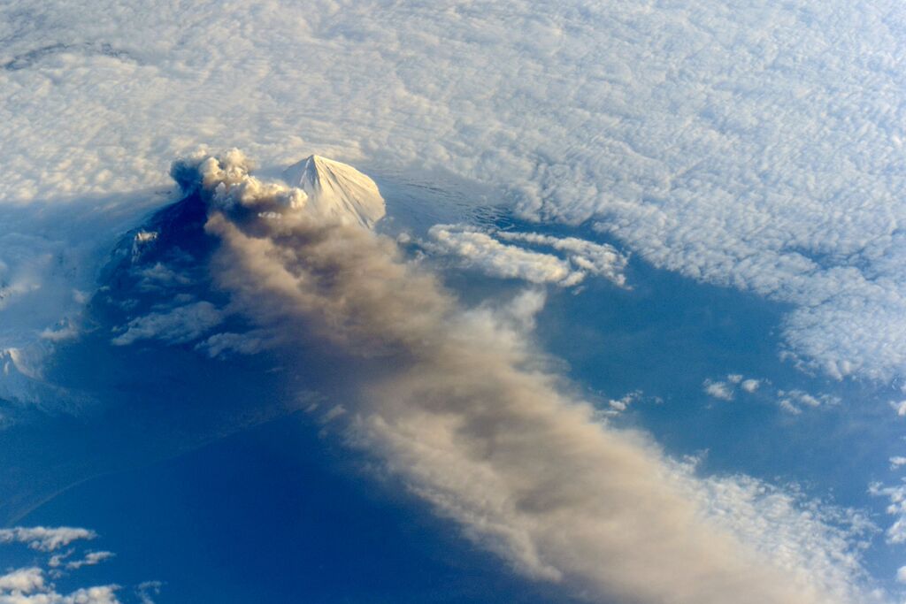 Zdjęcie przedstawia widziany z powietrza szczyt wulkanu, wypusszczający kłęby ciemnego dymu nad chmurami