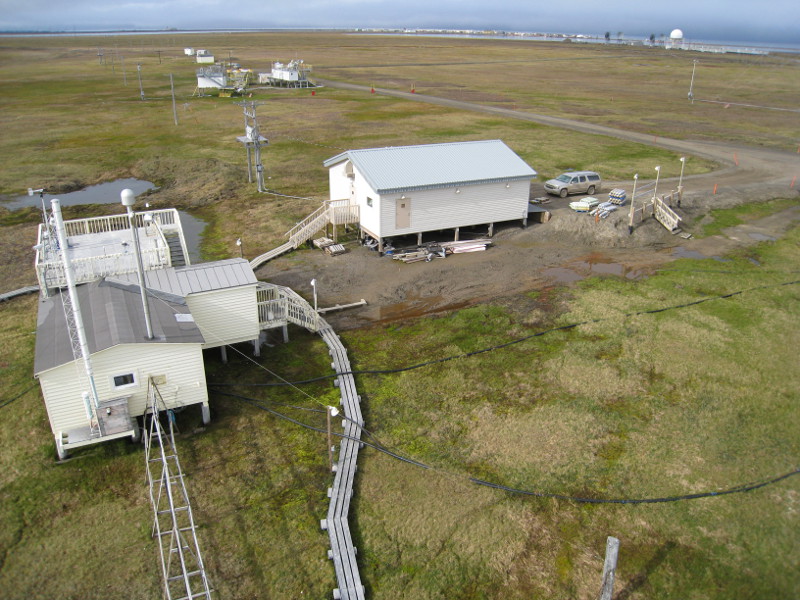 Zdjęcie przedstawia płaski obszar porośnięty rachityczną trawą i białe budynki z aparaturą naukową.