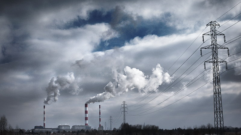 Zdjęcie przedstawia elektrownię z oddali, widoczne są trzy wysokie, cienkie kominy, dym mieszający się z chmurami i przewody elektryczne ze słupami po prawej stronie