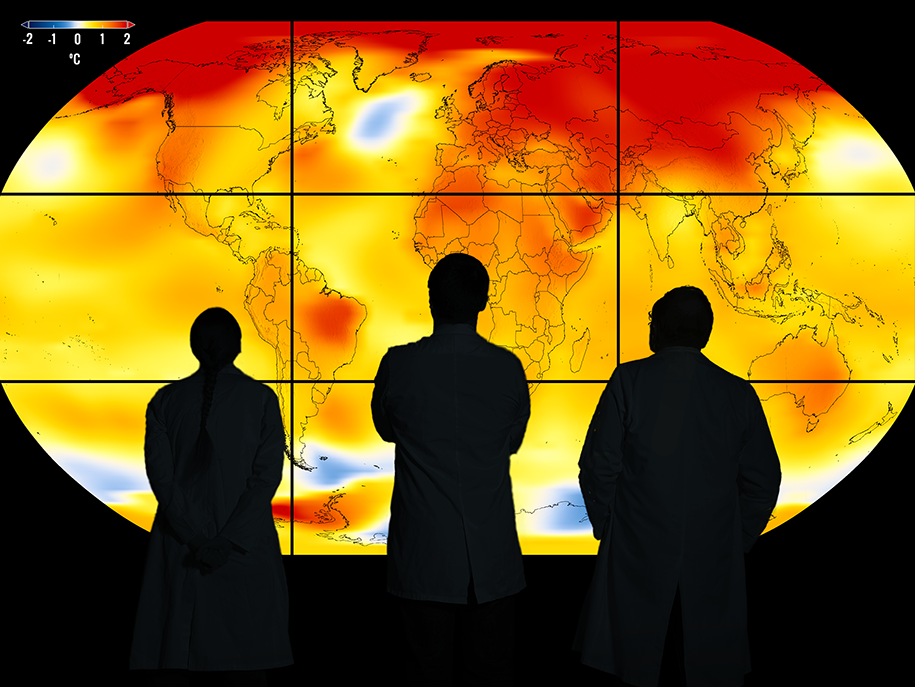 Zdjęcie przedstawia trzy ciemne syylwetki ludzi wpatrujących się w wyświetloną na dużym ekranie mapę świata w ciepłych kolorach