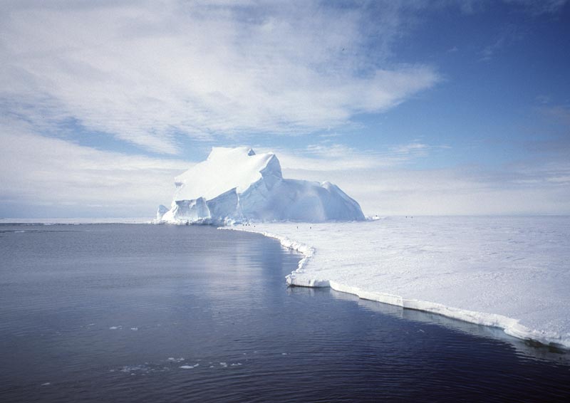 Zdjęcie przedstawia pogranicze między lodem a oceanem, widoczny jest duży płaski płat lodu z górą lodową w oddali oraz granatowa tafla oceanu.