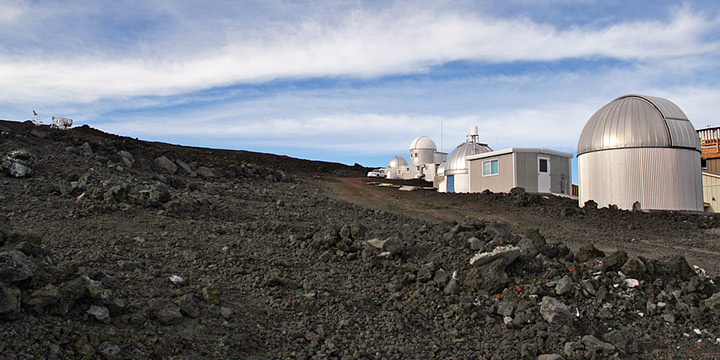 Zdjęcie przedstawia czarne zbocze wulkaniu i zabudowania obserwatorium z kopułkami itd.
