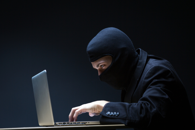 Zdjęcie przedstawia meżczyznę w kominiarsce stukającego w klawiaturę laptopa