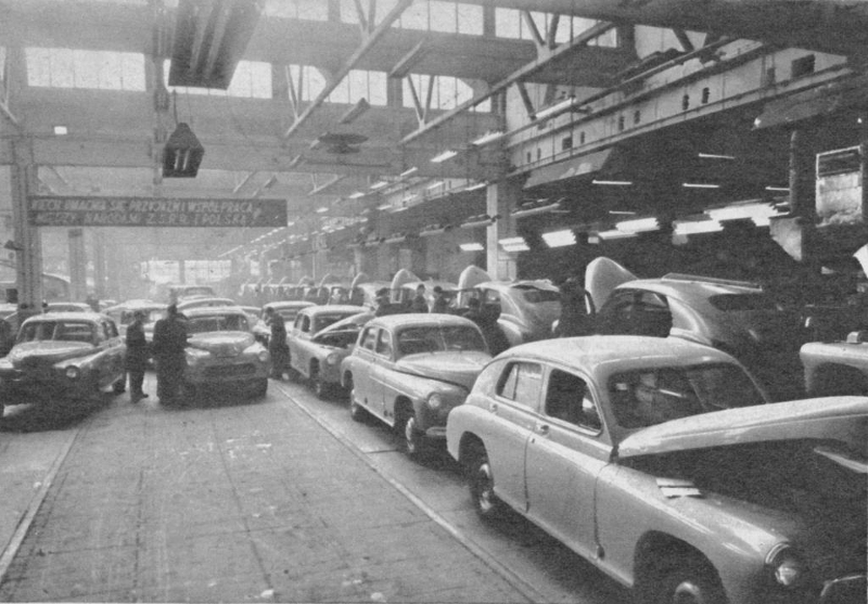 czarno białe zdjęcie przedstawia halę fabryczną z rzędem montowanych samochodów