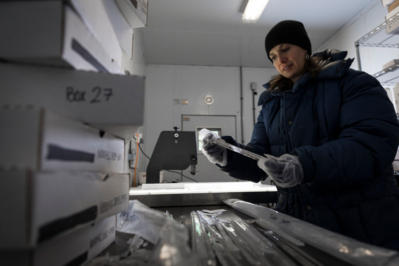 Zdjęcie przedstawia laboratorium z pudłami, w środku stoi kobieta w kurtce i czapce, na dłoniach ma ciepłe rękawiczki oraz platikowe rękawiczki ochronne, trzyma podłużną próbkę lodu - pręt długości ok. 40 cm.