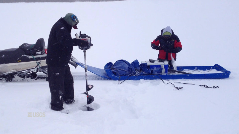 Zdjęcie: naukowcy pobierają próbki wody z zamarzniętej rzeki. Widać mężczyznę wiercącego w lodzie, w tle skuter śnieżny z doczepionymi z tyłu sankami. 