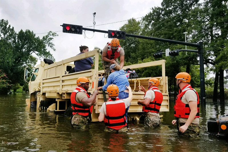 Zdjęcie przedstawia zalaną ulicę, po środku specjalny samochód z otwartym nadwoziem i na wysokich kołach. Grupa ludzi w czerwonych kamizelkach pomaga do niego wsiąść zwyczajnie ubranym osobom. 