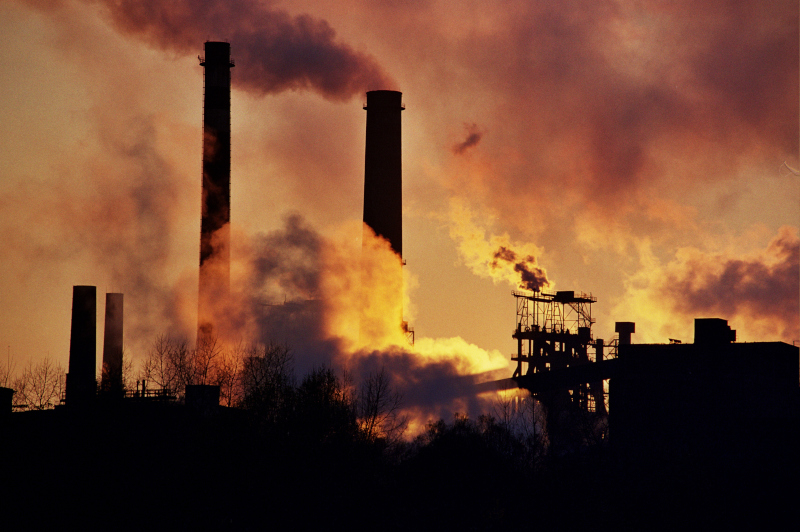 Zdjęcie przedstawia kominy fabryki na tle zachodu słońca, które emitują gęsty dym.
