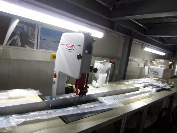 Zdjęcie: laboratorium badania rdzeni lodowych, na stołach leżą podłużne, zafoliowane cylindry, pośrodku stoi urządzenie do cięcia próbek