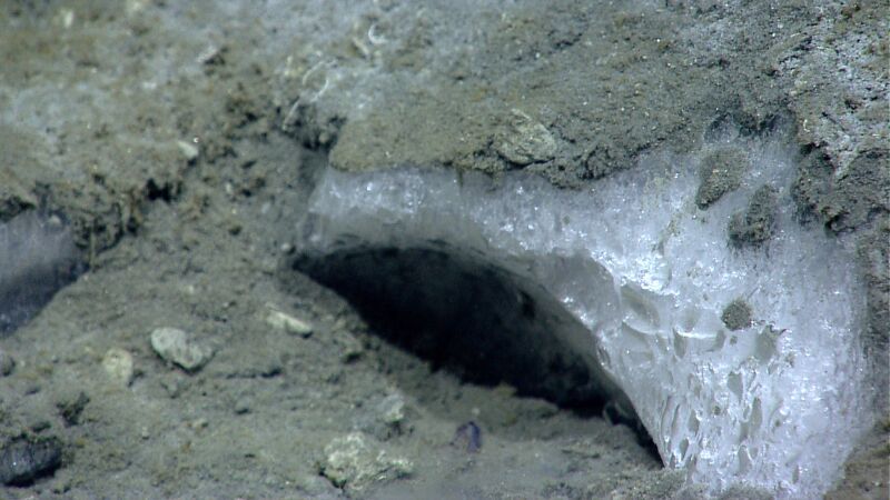 Zdjęcie: W niskich temperaturach i środowisku wodnym metan przyjmuje postać klatratów. Widoczny biały, matowy kryształ przysypany szarym piachem.