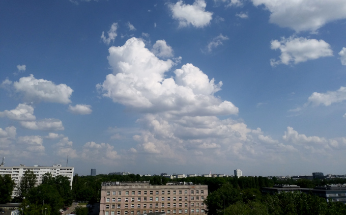 Zdjęcie: Drobne chmury konwekcyjne (cumulus) nad Warszawą, widać krajobraz miejski z wielopiętrowymi budynkami i drzewami, nad nim niebo z niewielkimi, białymi kłębiastymi chmurami.
