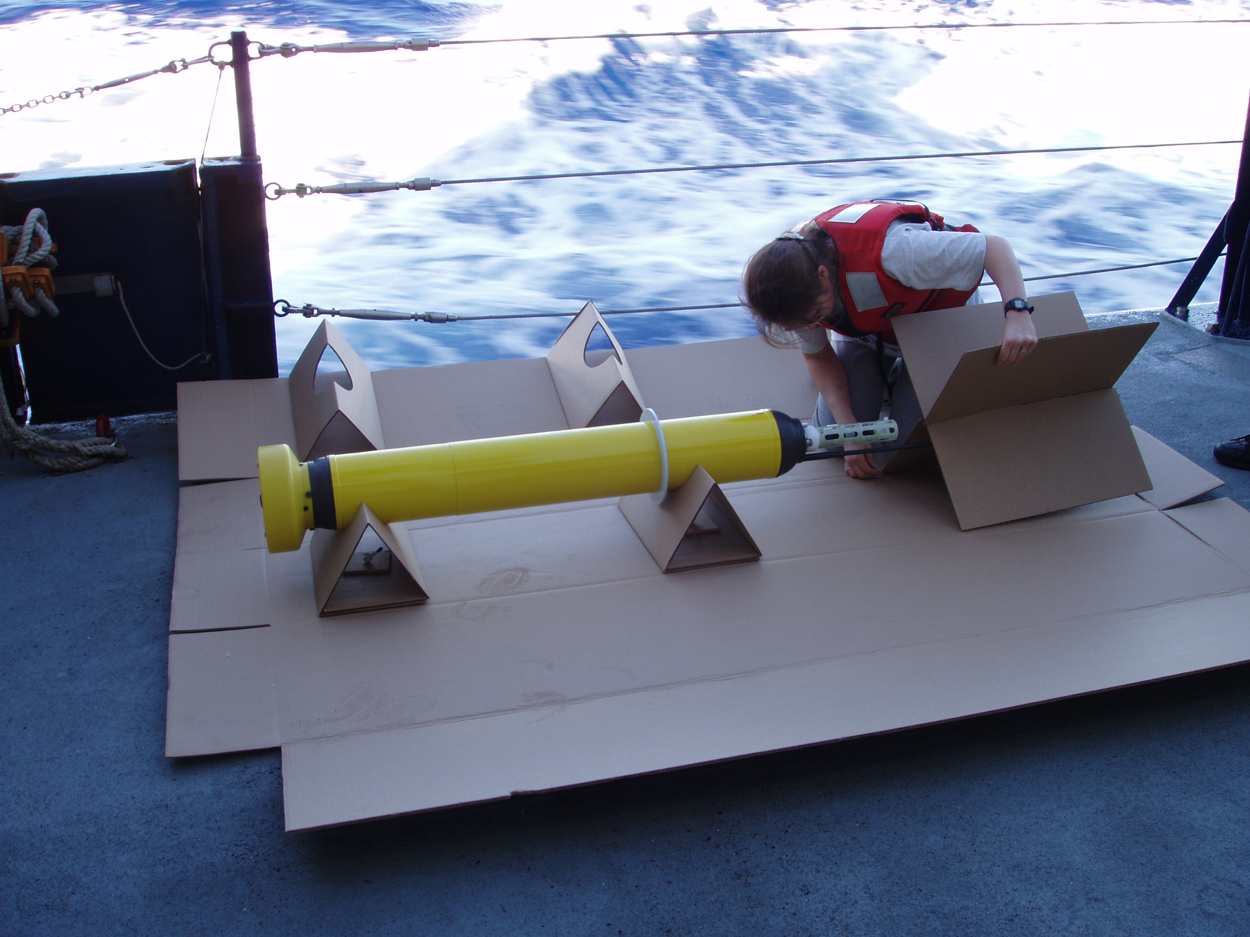 Zdjęcie: przygotowywanie pływaka Argo do umieszczenia w morzu. Na pokładzie statku leży ok. półtorametrowy, żółty cylinder z iglicą, nad nim pochyla się kobieta w kamizelce asekuracyjnej.