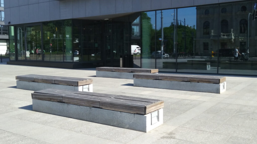 Zdjęcie dekoracyjne: ławki na betonowym dziedzińcu obok biurowca.