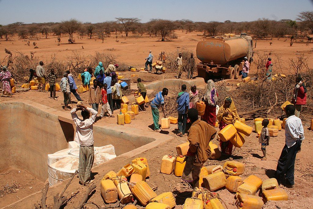 Zdjęcie: Dostawa wody podczas suszy w Afryce, 2010-2011. Widać wysuszony krajobraz, cysternę z wodą i dużą grupę ludzi z żółtymi plastikowymi kanistrami.