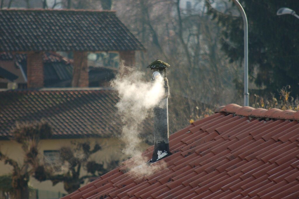  Zdjęcie ilustracyjne: dymiący komin na spadzistym dachu niewielkiego domu.