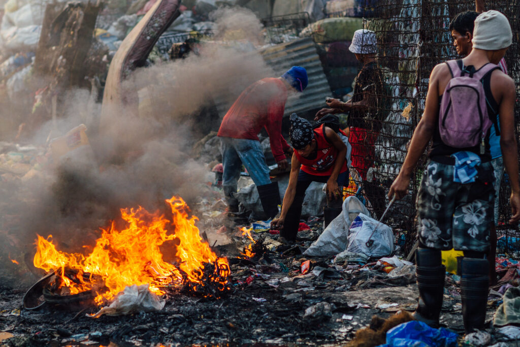 Zdjęcie przedstawiające grupę kilku mężczyzn na tle zwałów śmieci, na pierwszym planie ogień. 