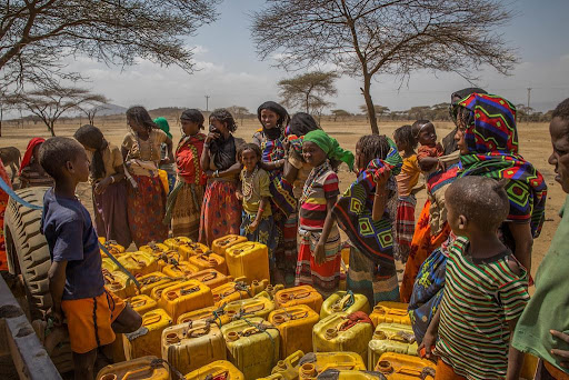 Obrazek przedstawia mieszkanki i mieszkańców Afryki, którzy stoją dookoła plastikowych baniaków z wodą. W tle widać sawannę.