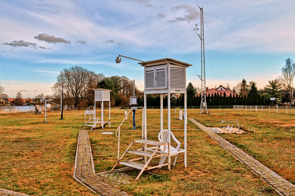 Zdjęcie: stacja meteorologiczna (sieć pomiarowa IMGW). Na krótko przystrzyżonym trawniku, na wysokich nóżkach stoją klatki pomiarowe (skrzynki z instrumentami), w tle także maszt. 