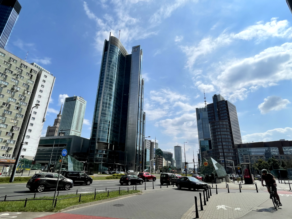 Zdjęcie: upalny dzień w Warszawie, Rondo ONZ, widać wysokie budynki, samochody i rowerzystę. 