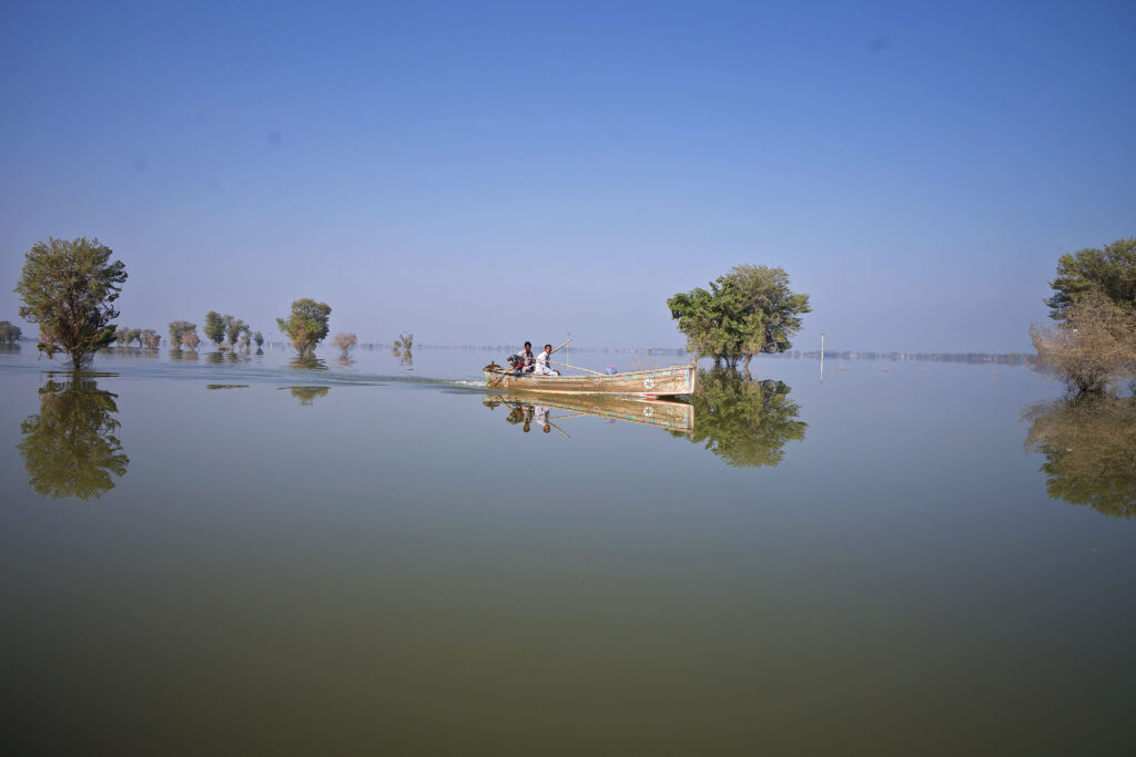 Powodzie 2022: zdjęcie z prowincji Sindh w Pakistanie podczas powodzi. Widać wielki obszar wody, z której wystają drzewa i po której dwóch ludzi płynie łodzią. 