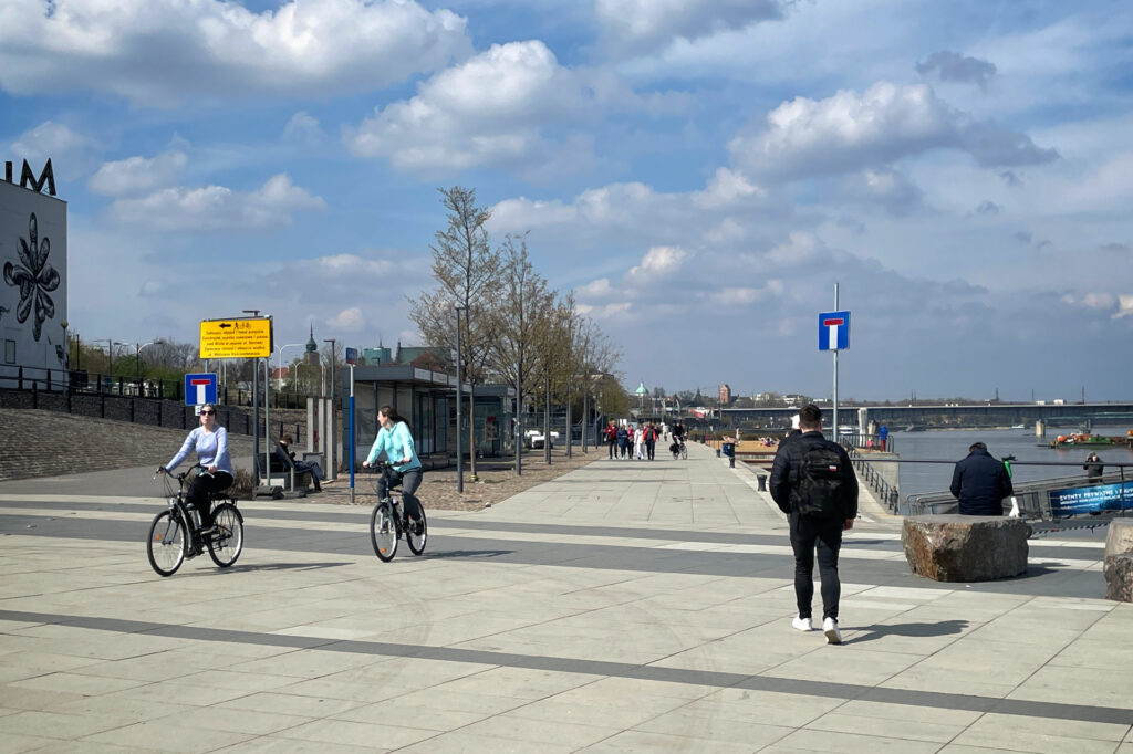 Zdjęcie: bulwary nad Wisłą w Warszawie. Wiosna, kwiecień 2023. Zalana słońcem aleja ze spacerowiczami i rowerzystami. 