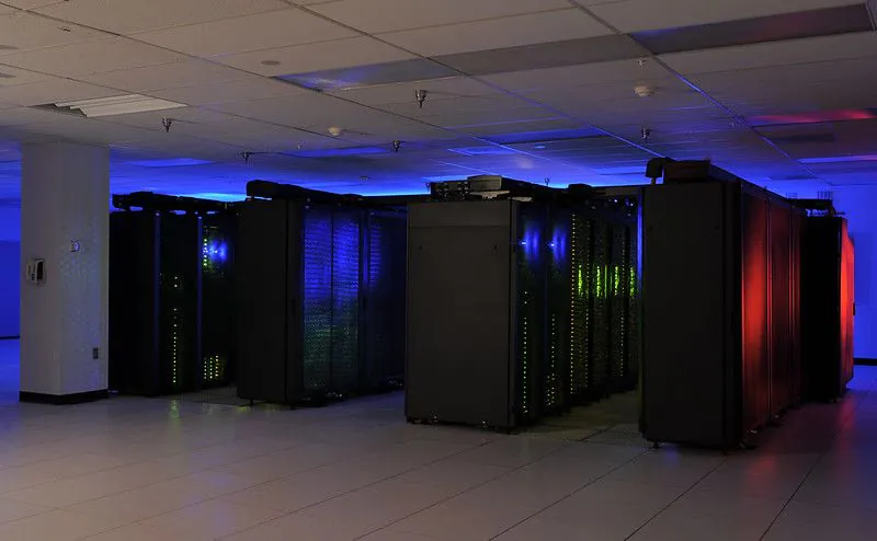 Zdjęcie: superkomputer NASA, widać ciemne pomieszczenie wypełnione czarnymi szafami, rozświetlanymi przez niebieskie, zielone i czerwone światełka. 
