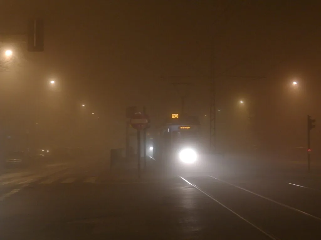 Zdjęcie przedstawiające smog w Krakowie: ulica i przystanek tramwajowy z tramwajem, po zmroku, zasnute brudną mgłą.