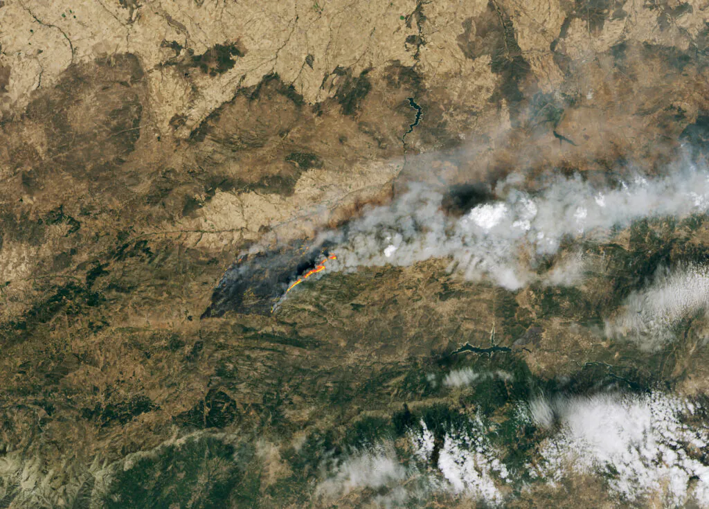 Zdjęcie satelitarne pokazujące pożary w Hiszpanii, widać wysuszoną powierzchnię Ziemi, cienki pas pożarów, unoszące się w atmosferze obłoki dymu.