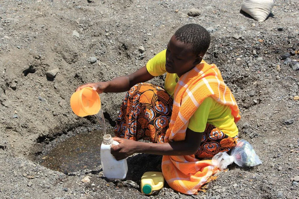 Zdjęcie: skutki zmiany klimatu. Kolorowo ubrana, czarna kobieta wydobywa resztki wody z wyschniętego koryta rzeki. Plastikową miseczką nabiera wody z niewielkiej kałuży do butelki. 
