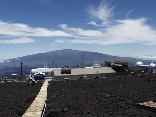 Zdjęcie: obserwatorium Mauna Loa, widoczne niskie, prostopadłościenne budynki i półsferyczne kopułki na tle góry, na pierwszym planie kamieniste zbocze.
