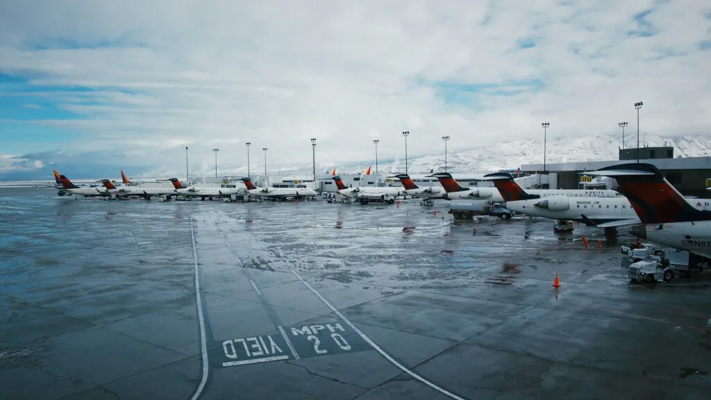 Zdjęcie: rząd samolotów przy terminalu dużego lotniska, w tle ośnieżone góry.