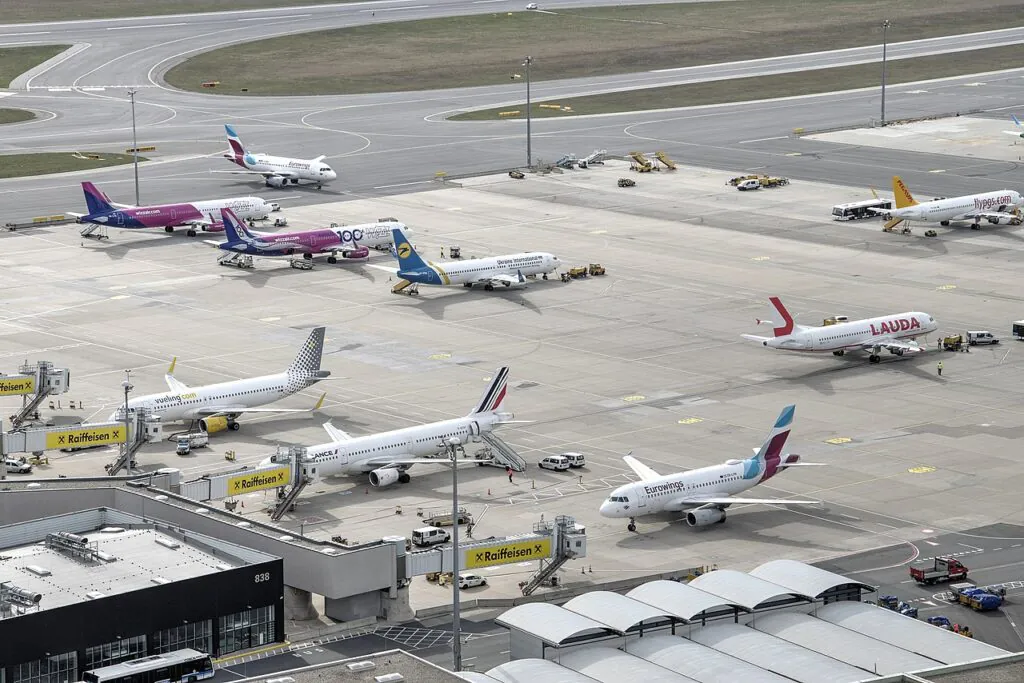 Zdjęcie: płyta lotniska z zaparkowanymi wieloma samolotami pasażerskimi. 