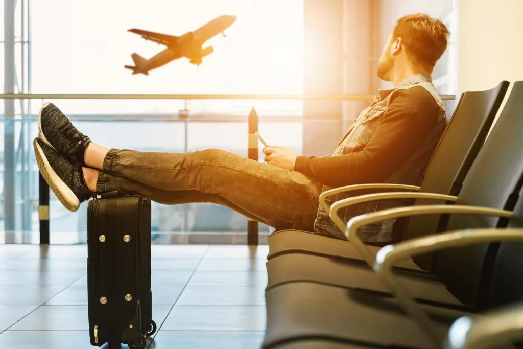 Lotnictwo a klimat. Zdjęcie: Młody mężczyzna w poczekalni na lotnisku spogląda za okno, za którym widać startujący samolot.