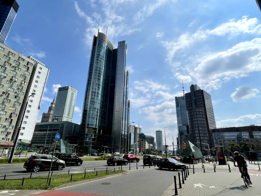 Zdjęcie: upalny dzień w Warszawie, Rondo ONZ, widać wysokie budynki, samochody i rowerzystę. 