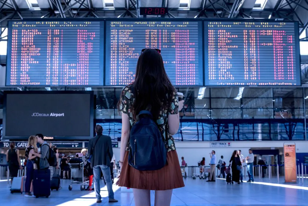 Zdjęcie: mloda dziewczyna w hali odlotów na lotnisku, patrzy na tablice z rozkładem lotów i bramek. 