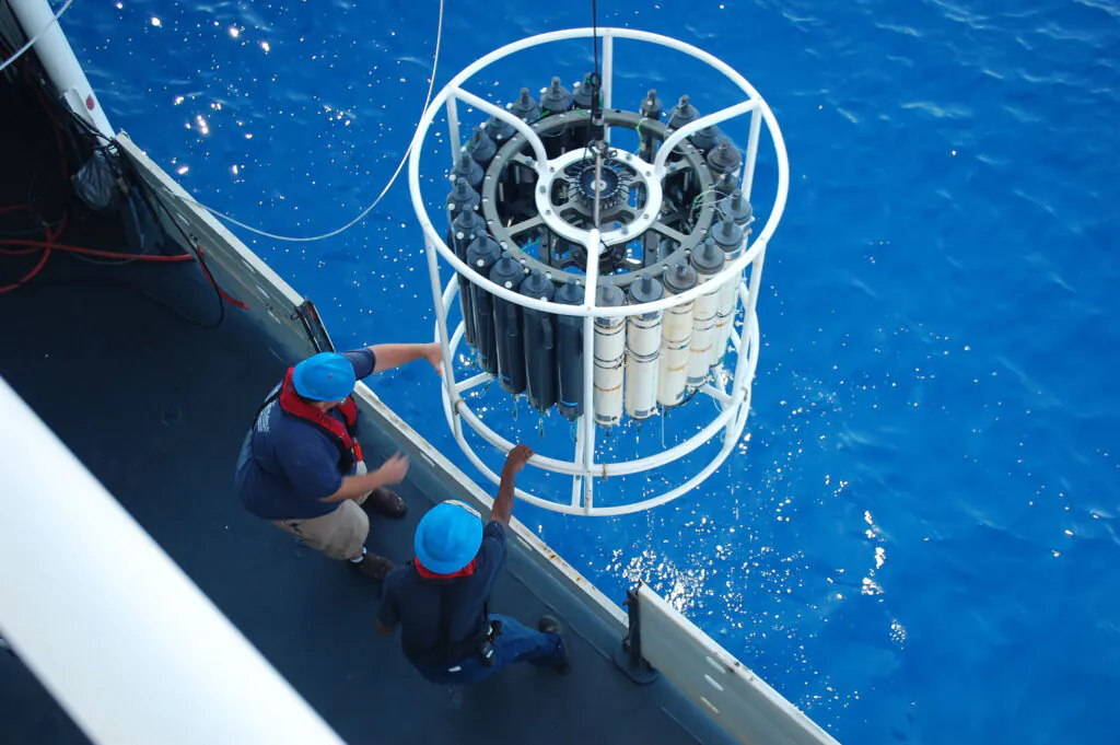 Zdjęcie: widziany z góry fragment statku, na którym stoi dwóch ludzi w niebieskich kaskach, za burtą zwisa wielki ażurowy cylinder z przyczepionymi cylindrycznymi pojemnikami z instrumentami. 