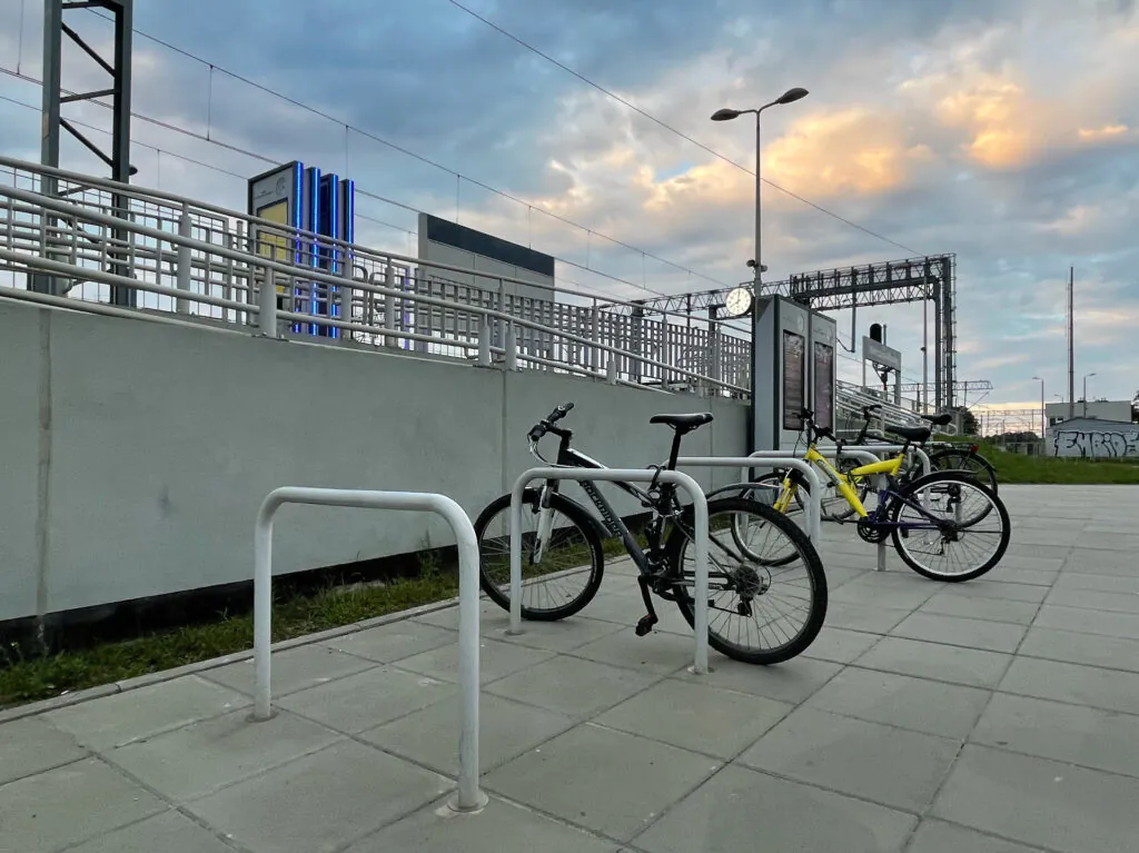 Zdjęcie: stojaki na rowery (z przyczepionymi do nich rowerami) przy peronie niewielkiej stacji kolejowej.  