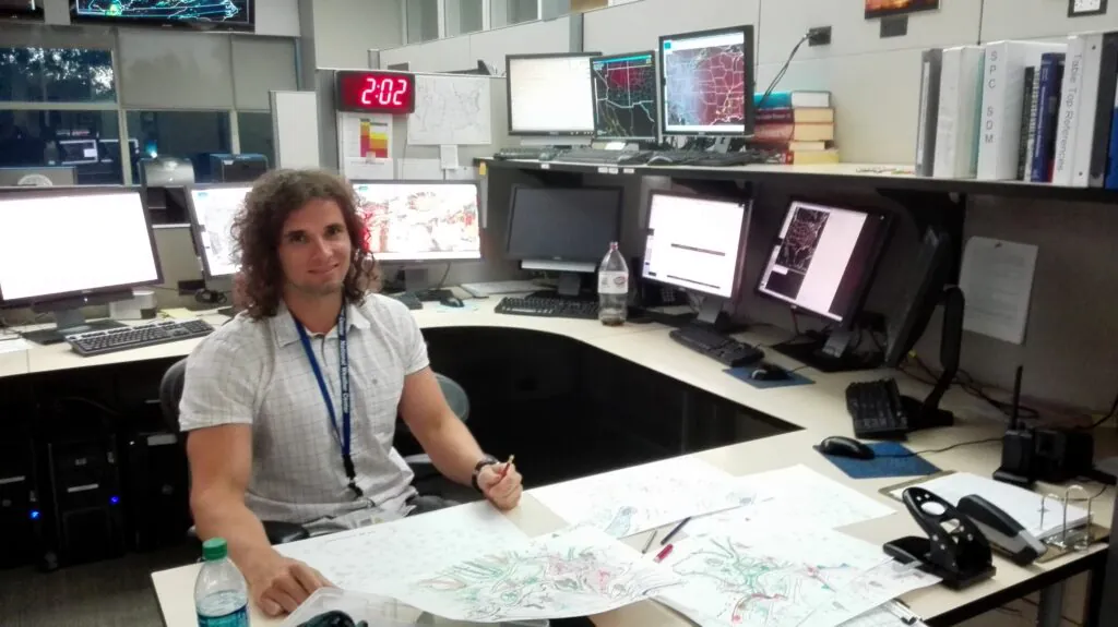 Zdjęcie: Mateusz Taszarek w biurze, otoczony ekranami wyświetlającymi dane meteorologiczne i przeglądający mapę pogody.