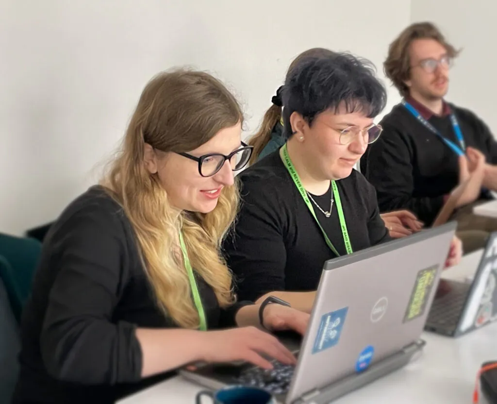 Zdjęcie: Alicja Dec i Anna Sowa podczas warsztatów. Uśmiechnięte siedzą przy stoliku, każda ze swoim laptopem.