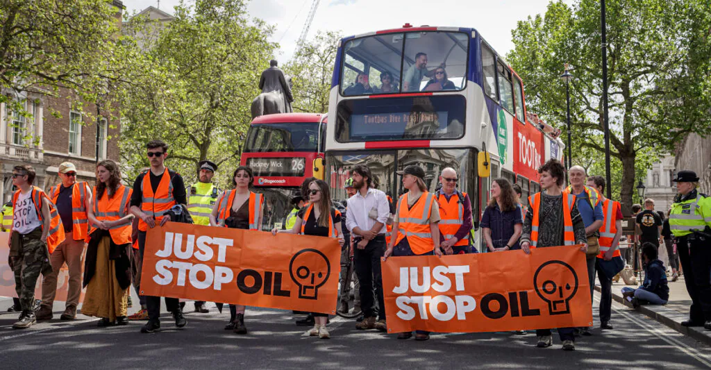 Zdjęcie ilustracyjne: protest "Just STop Oil". Grupa ludzi w jaskrawych kamizelkach i z transparentami "Just stop oil" stoi w poprzek ulicy. Za ich plecami widać policjantów i dwa piętrowe autobusy.