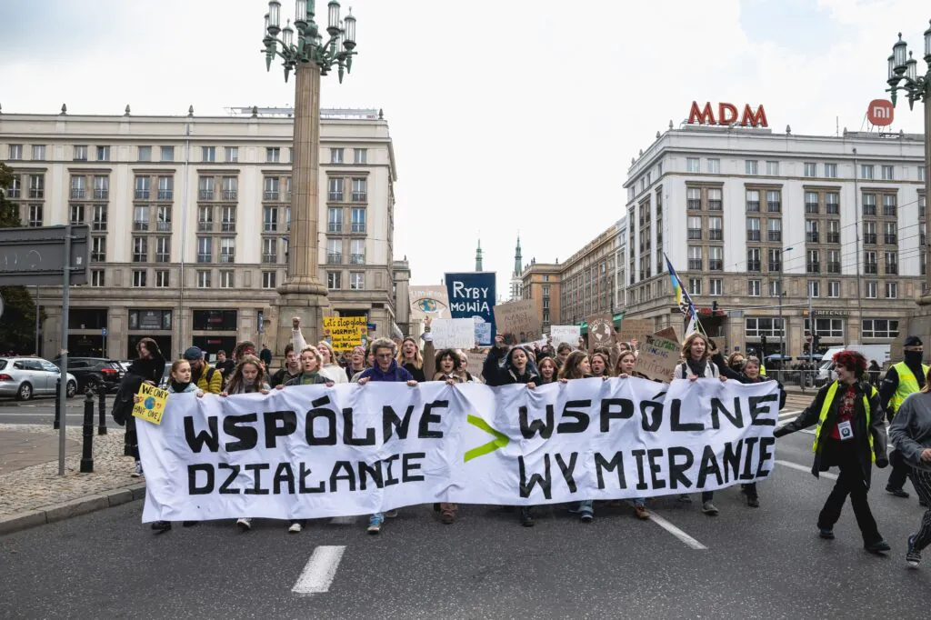 Zdjęcie ilustracyjne: marsz klimatyczny w Warszawie. Grupa ludzi maszeruje ulicą z transparentem "wspólne działanie > wspólne wymieranie". 