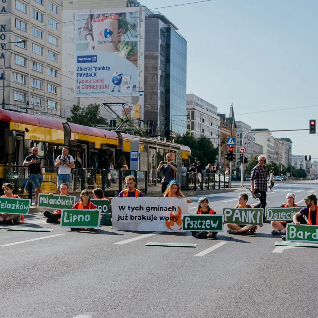 Zdjęcie ilustracyjne: protest "Ostatniego Pokolenia". Grupa ludzi siedzi w poprzek wielopasmowej ulicy, trzymając tablice z nazwami miejscowości oraz tablicę z napisem "W tych gminach brakuje wody". 