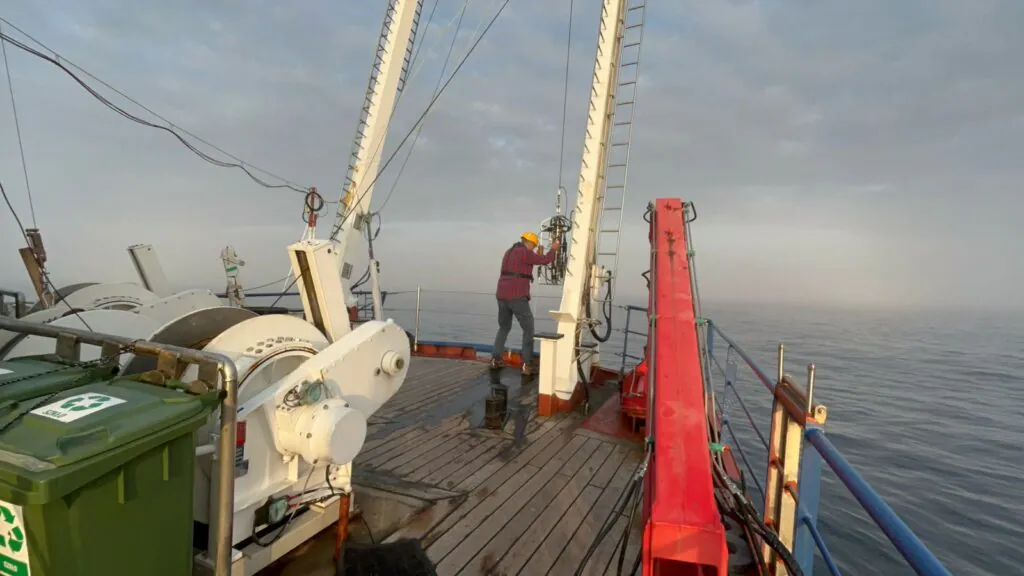 Zdjęcie: badania na pokładzie RV Oceania. Widać naukowca opuszczającego do wody przyrządy do zbierania próbek za pomocą windy na rufie statku.