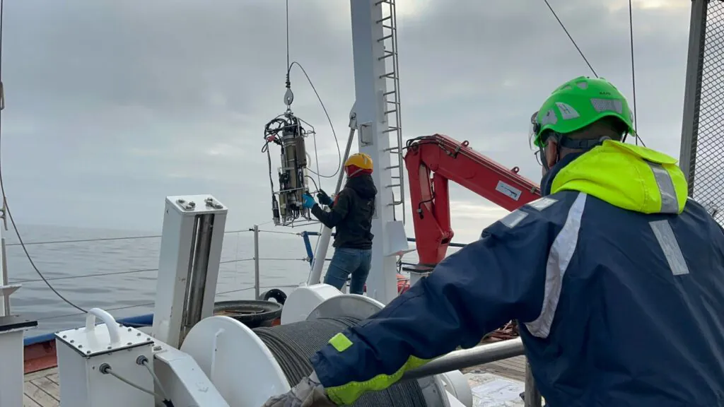 Zdjęcie: badania na pokładzie RV Oceania. Widać naukowca i naukowczynię opuszczających do wody przyrządy do zbierania próbek za pomocą windy na rufie statku.